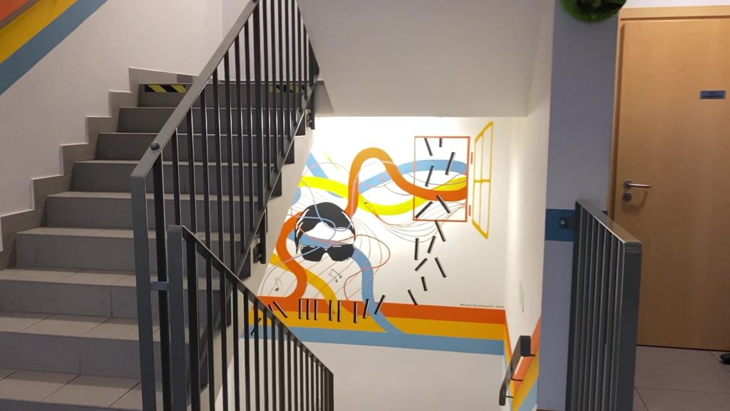 Zdjęcie przedstawia szkolny korytarz, widok na klatkę schodową i kolorowy mural.