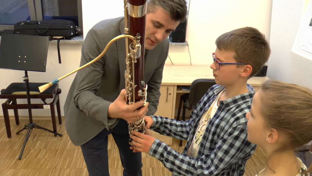 Zdjęcie przedstawia nauczyciela fagotu prezentującego instrument dwójce kandydatów do szkoły muzycznej.
